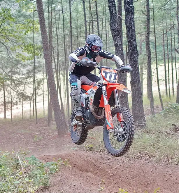 Sam jumping his orange KTM on a trail through pine woods