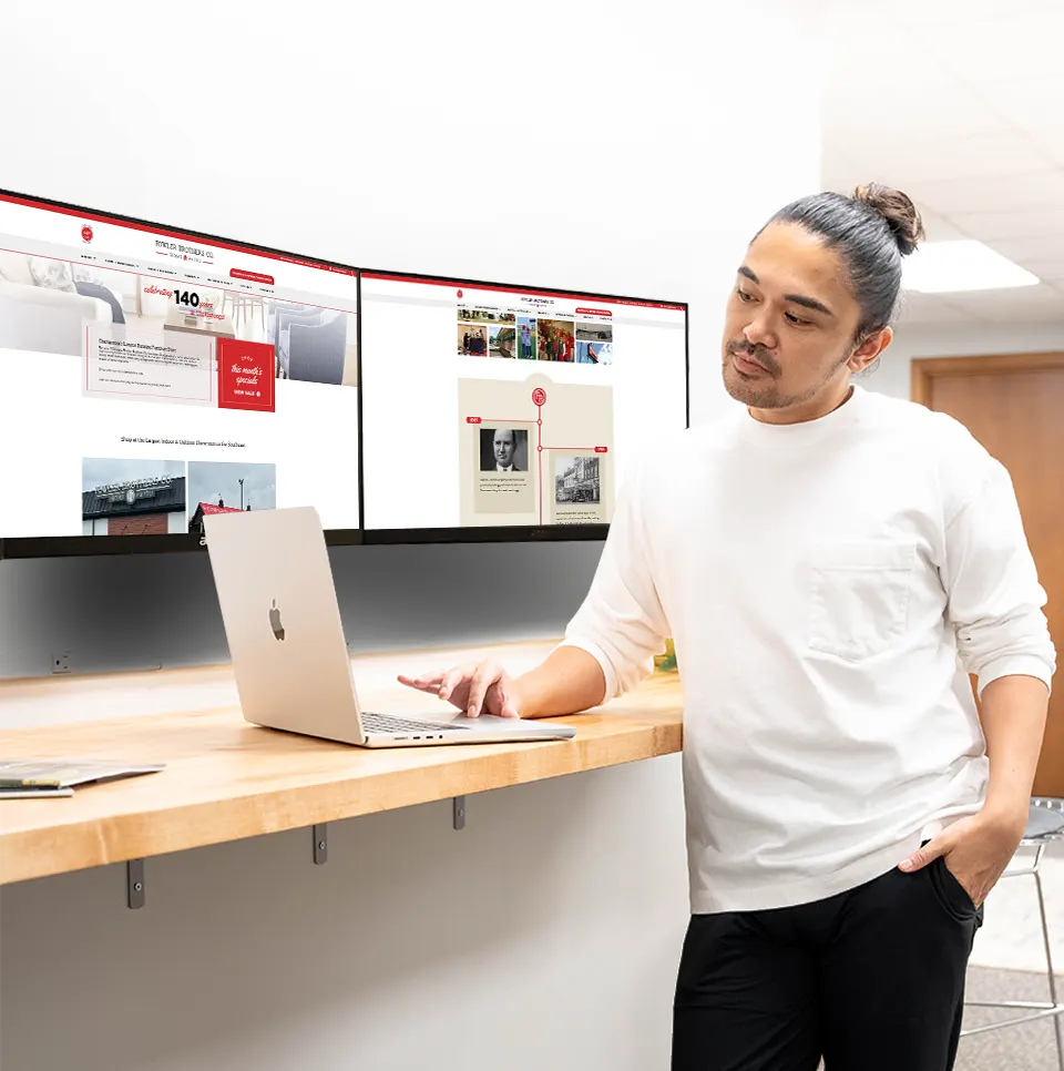 A Spectruss team member at a standing desk with the Fowler Brothers website on two monitors